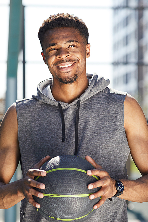 Waist up portrait of muscular African man holding basketball ball outdoors and smiling at camera , copy space