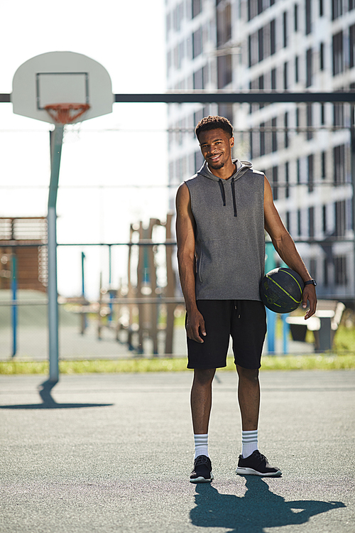 Full length portrait of African basketball player standing by hoop in outdoor court and smiling at camera, copy space