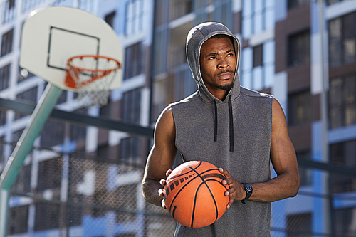 Waist up portrait of handsome African man holding ball while posing in basketball court outdoors, copy space