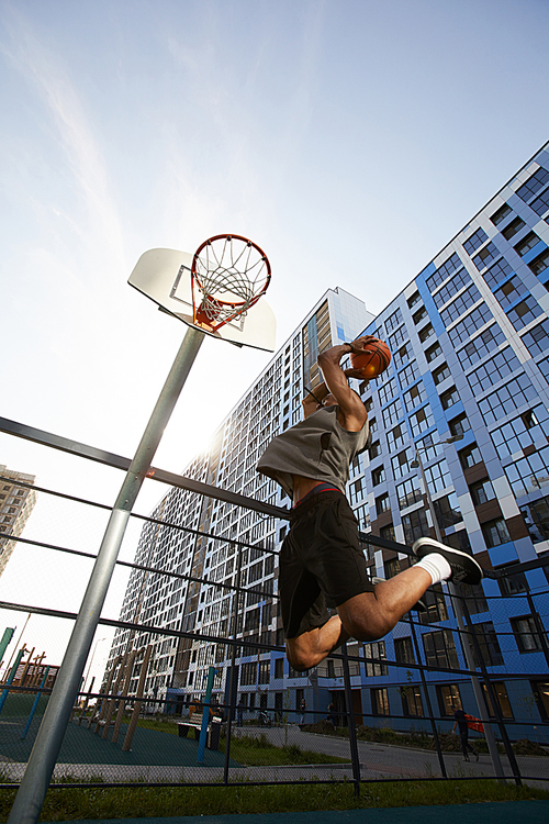 Low angle action shot of African basketball player jumping while shooting slam dunk in outdoor court, copy space