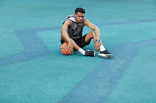 Wide angle portrait of handsome African sportsman sitting on floor in basketball court and , copy space