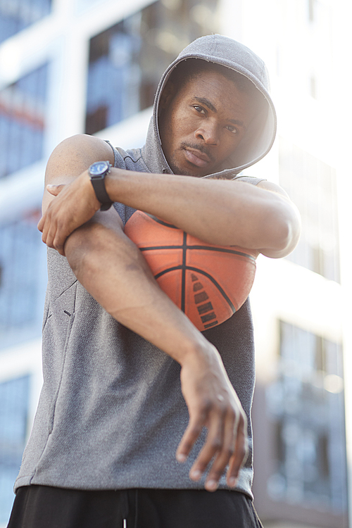 Waist up portrait of contemporary African man wearing hoodie posing with basketball ball in urban setting