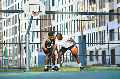 Wide angle action shot of two African-American guys playing basketball in outdoor court in urban setting, copy space