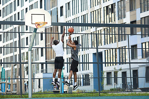Wide angle action shot of two African-American guys jumping while playing basketball in outdoor court, copy space