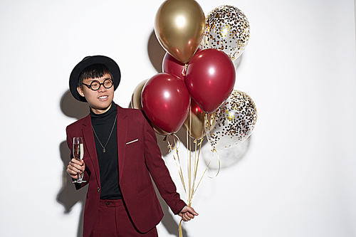 Waist up portrait of fashionable Asian man wearing maroon suit and stylish hat posing with balloons against white background at party, shot with flash, copy space