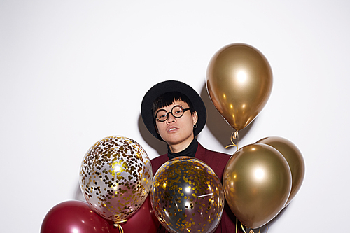 Waist up portrait of trendy Asian man holding bunch of golden balloons while posing against white background at party, shot with flash, copy space