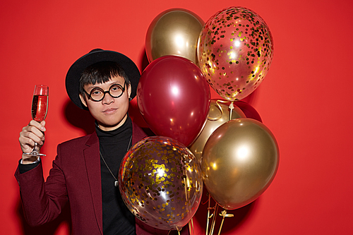 Waist up portrait of trendy Asian man holding bunch of golden balloons while posing against red background at party, shot with flash