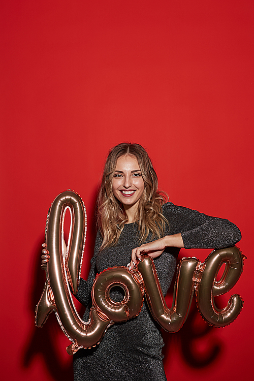 Waist up portrait of smiling young woman holding golden LOVE balloon while posing against red background at party, copy space