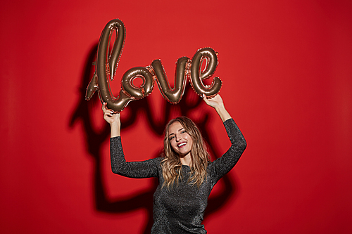 Waist up portrait of excited young woman holding golden LOVE balloon while standing against red background at party, copy space