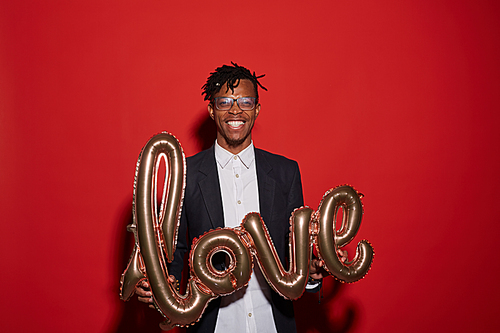Waist up portrait of contemporary African man holding golden LOVE balloon while standing against red background at party, copy space