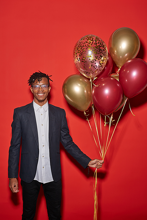 Portrait of smiling African man holding bunch of golden balloons while standing against red background at party, copy space