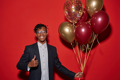 Waist up portrait of smiling African man showing thumbs up and holding bunch of golden balloons while standing against red background at party, copy space