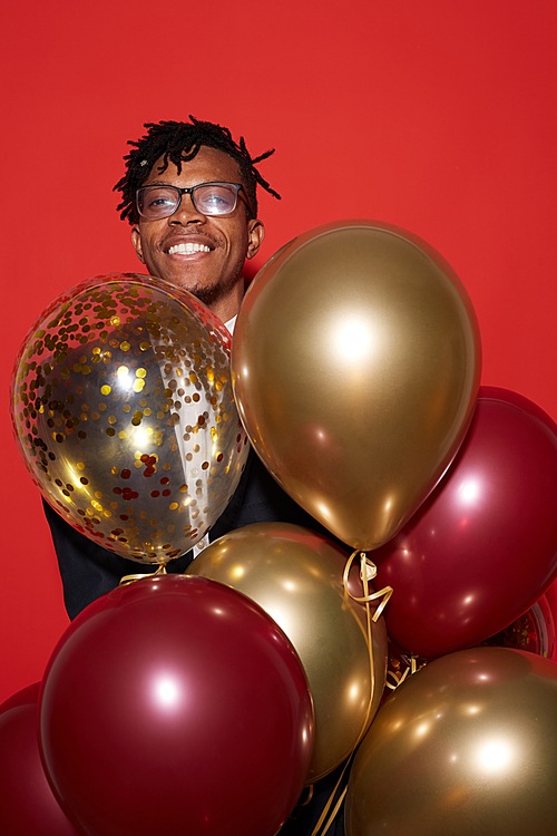 Waist up portrait of smiling African man holding bunch of golden balloons while standing against red background at party