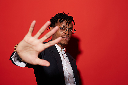 Waist up portrait of contemporary African man stretching hand to camera while standing against red background, shot with flash