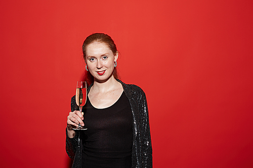 Waist up portrait of red haired young woman smiling at camera and holding champagne glass while posing against red background at party, copy space