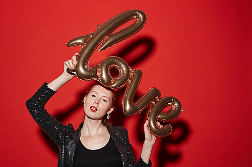 Waist up portrait of red haired young woman holding golden LOVE balloon over head while posing against red background at party, copy space