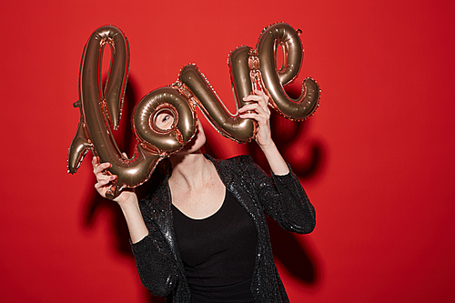 Waist up portrait of young woman holding golden LOVE balloon while posing against red background at party, shot with flash