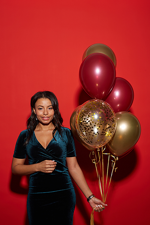 Waist up portrait of elegant African-American woman holding golden balloons while posing against red background at party, shot with flash