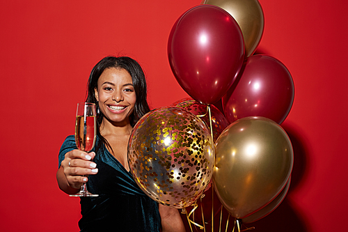 Waist up portrait of smiling African-American woman holding champagne glass and golden balloons while enjoying party, shot with flash on red background