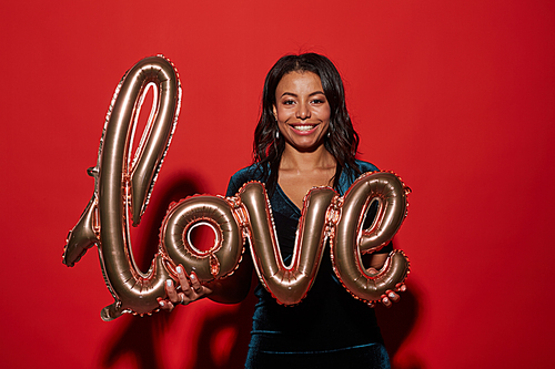 Waist up portrait of smiling African-American woman holding golden LOVE balloon while enjoying party, shot with flash on red background