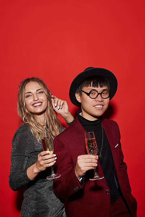 Waist up portrait of trendy young people holding champagne glasses while enjoying party, shot with flash on red background