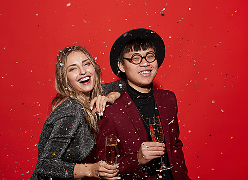 Waist up portrait of trendy young couple holding champagne glasses and smiling happily while enjoying party, shot with flash on red background