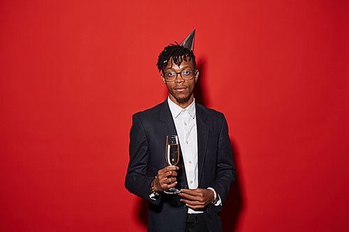 Waist up portrait of trendy African-American man holding champagne glass while posing against red background at party, shot with flash