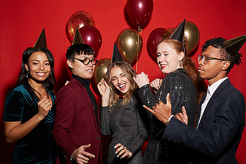 Multi-ethnic group of young people dancing together over red background with party balloons, copy space