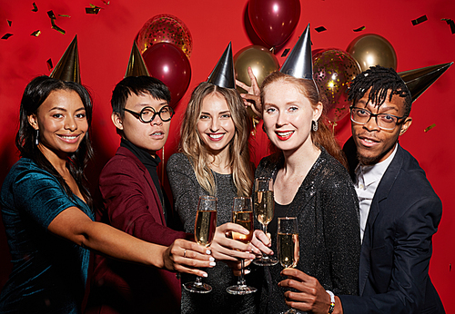 Multi-ethnic group of young people clinking champagne glasses while posing over red background enjoying party, shot with flash