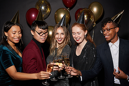 Multi-ethnic group of young people clinking champagne glasses while posing over black background enjoying party, shot with flash