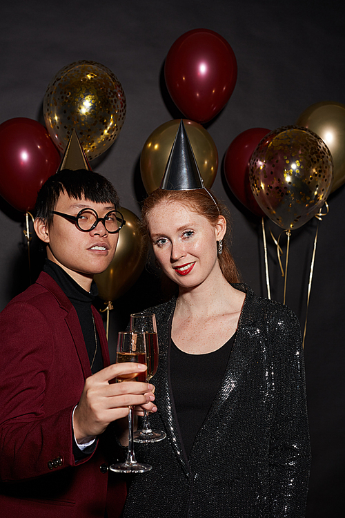 Waist up portrait of young couple holding champagne glasses while posing over black background enjoying party, shot with flash