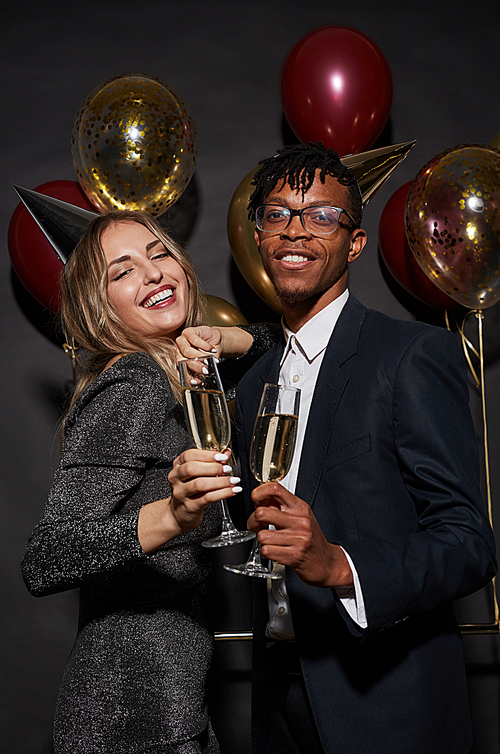Waist up portrait of mixed race young couple holding champagne glasses while posing over black background enjoying party, shot with flash