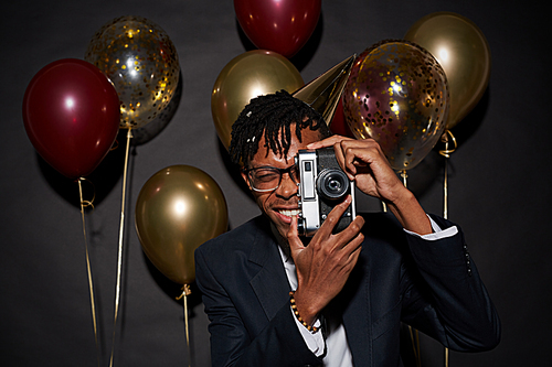 Waist up portrait of contemporary African man holding photo camera while posing against black background with party balloons, shot with flash