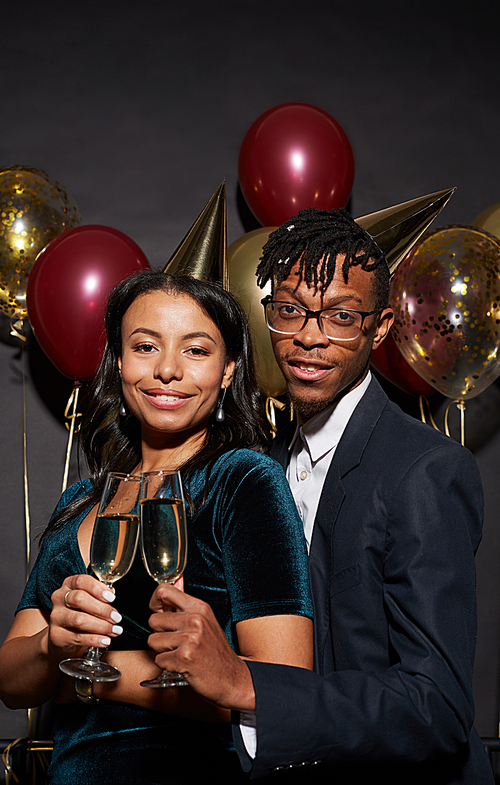 Waist up portrait of elegant mixed race young couple holding champagne glasses while posing over black background t party, shot with flash