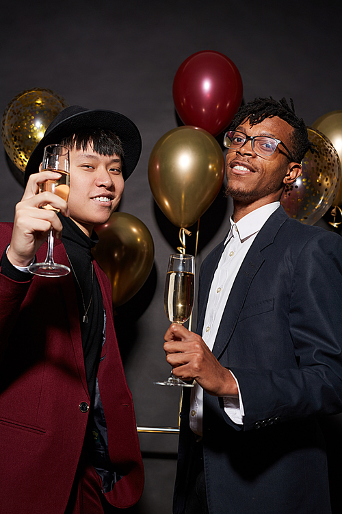 Portrait of two ethnic young men raising glasses to camera while posing against black background at party, shot with flash