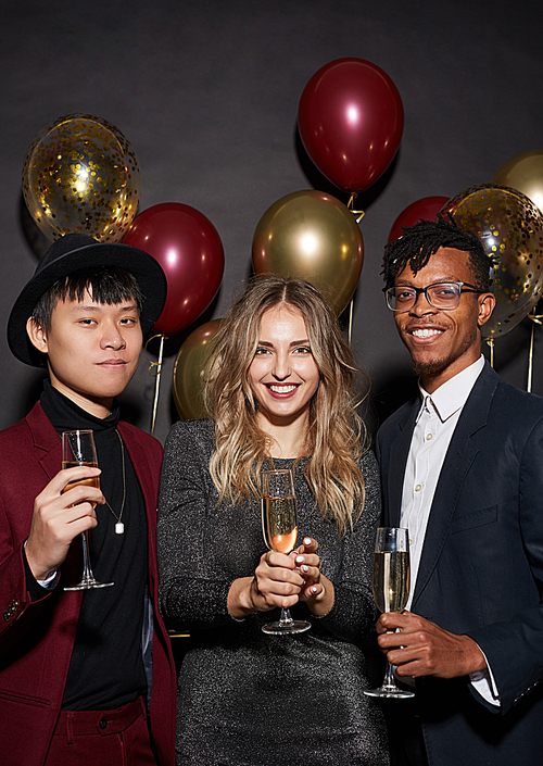 Group of contemporary young people holding champagne glasses and smiling cheerfully while posing against black background at party, shot with flash