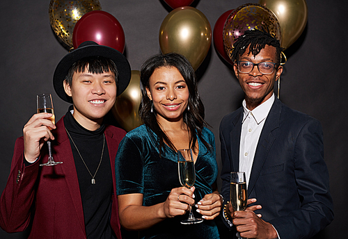 Group of ethnic young people holding champagne glasses and smiling happily while posing against black background at party, shot with flash