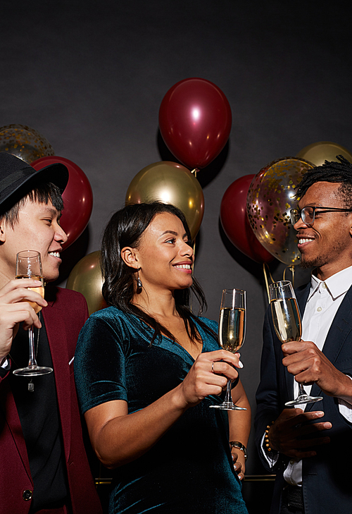 Group of ethnic young people holding champagne glasses standing against black background enjoying party, shot with flash