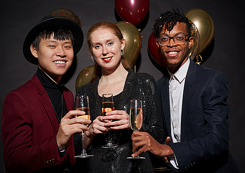 Group of elegant young people holding champagne glasses and smiling happily while posing against black background at party, shot with flash