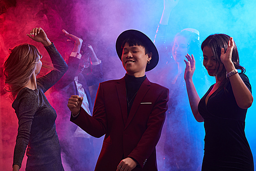 Waist up portrait of trendy Asian man dancing in smoky nightclub surrounded by beautiful women, copy space