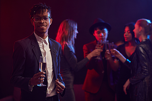 Waist up portrait of trendy African man holding champagne glass while posing at party in night club with people in background, copy space
