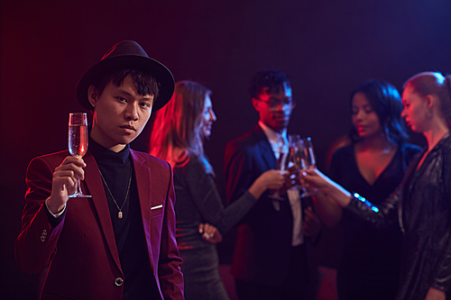 Waist up portrait of trendy Asian man holding champagne glass while posing at party in night club with people in background, copy space