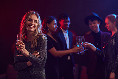 Waist up portrait of beautiful young woman holding champagne glass while posing at party in night club with people in background, copy space