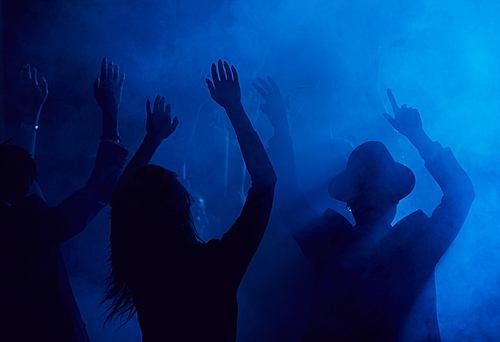 Silhouettes of young people jumping and raising hands while enjoying party in smoky nightclub lit by blue light, copy space