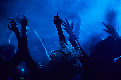 Silhouettes of young people jumping and raising hands while enjoying music concert in smoky nightclub lit by blue light, copy space