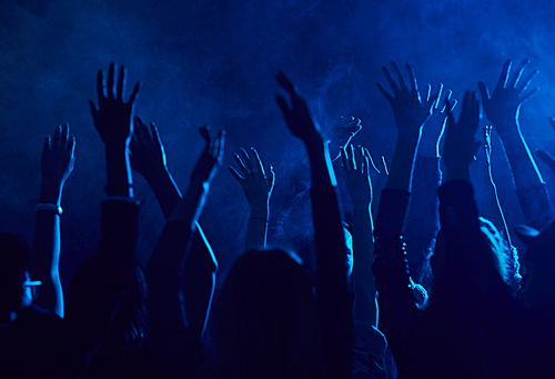 Silhouette of large group of people raising hands while enjoying music concert in smoky nightclub lit by blue light, copy space
