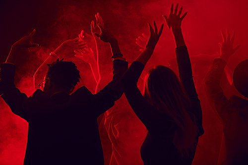 Dark silhouettes of young people dancing and raising hands while enjoying party in smoky nightclub lit by red light, copy space