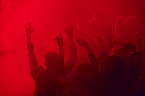 Silhouettes of large group of people dancing and raising hands while enjoying party in smoky nightclub lit by red light, copy space