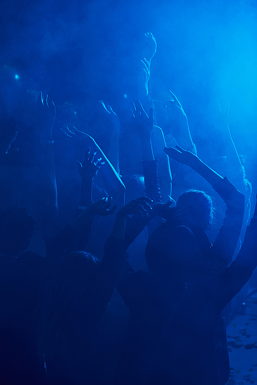Silhouettes of large group of people dancing and raising hands while enjoying party in smoky nightclub lit by blue light, copy space