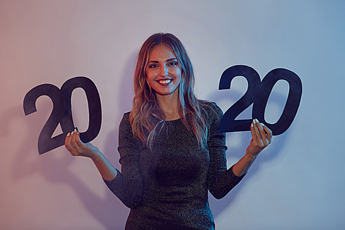 Waist up portrait of beautiful young woman holding 2020 sign during Christmas party in nightclub